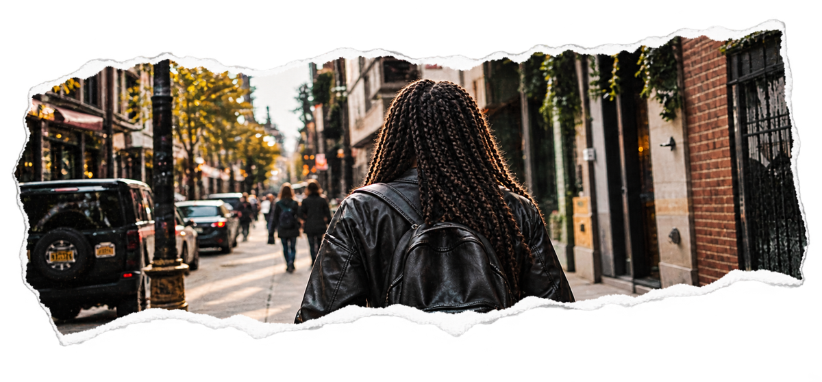 Someone with long braids and a leather backpack walking down a busy city street, seen from behind.